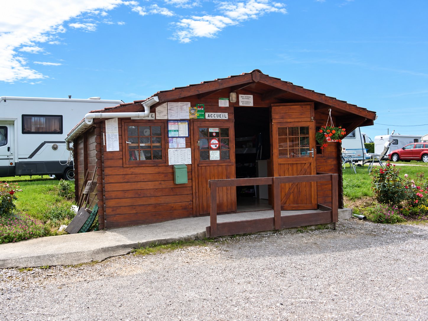 Chalet en bois de l’accueil du camping, avec des affichettes sur la façade, des fleurs suspendues et des camping-cars derrière