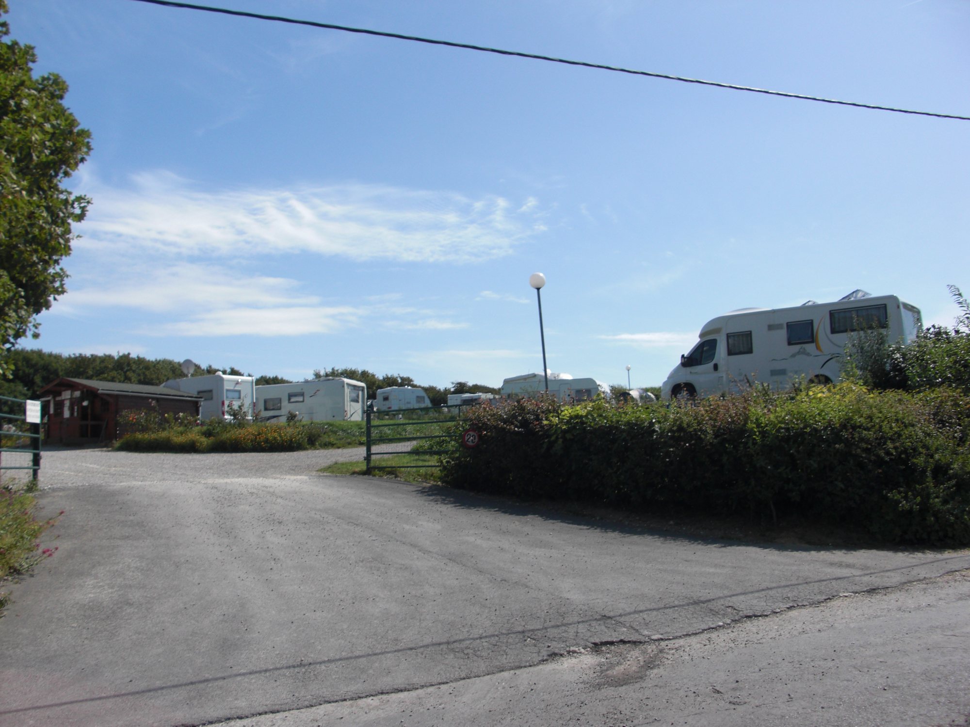 Entrée du camping bordée d’une haie, avec un camping-car, des caravanes et un petit bâtiment en bois sous un ciel bleu