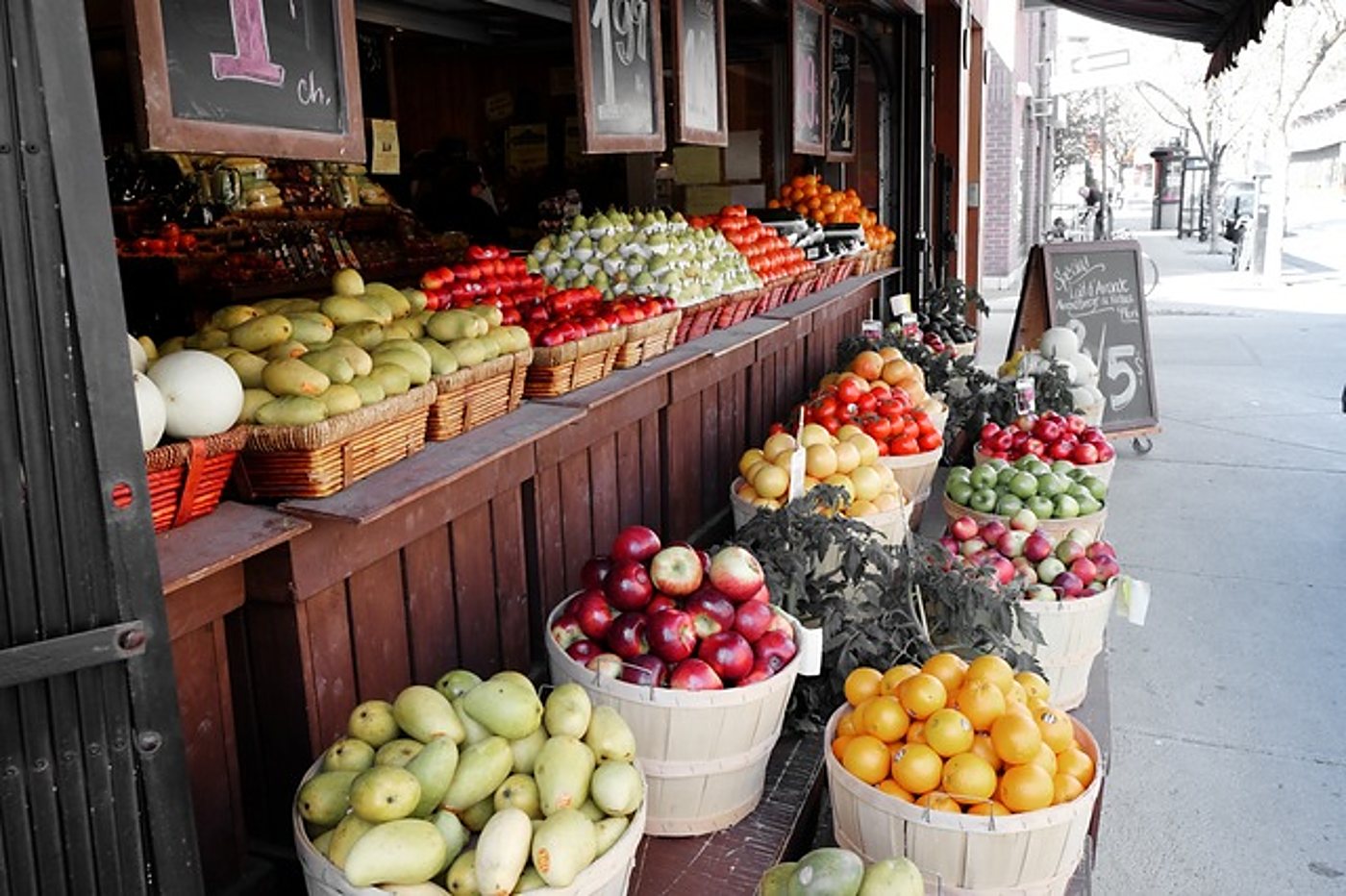Étal de bois garni de paniers de fruits frais devant une épicerie, avec des ardoises de prix